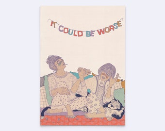 May include: A rectangular art print featuring a line drawing of two individuals sitting together on a patterned surface with a small cat. Above them, a colorful banner displays the text IT COULD BE WORSE in multi-colored block letters.