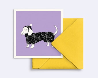 May include: A square greeting card featuring a line art illustration of a dachshund wearing a black and white patterned onesie with a matching hood and pom-pom, set against a solid lavender background, accompanied by a bright yellow envelope.
