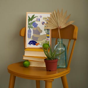 May include: A framed illustration of a person wearing a blue swimsuit and a blue face mask, sitting in a room with plants and a window. The illustration is on a yellow chair with a stack of books, a potted plant, and a glass bottle.