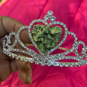 May include: A silver tiara with a heart-shaped green stone in the center. The tiara is decorated with rhinestones.