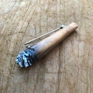 Puede incluir: Un pin de madera con forma de cigarrillo con un cierre de plata. El cigarrillo está pintado para que parezca que está encendido y tiene ceniza gris.