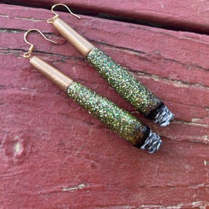 May include: A pair of handmade earrings designed to resemble rolled cigarettes. The earrings feature gold-colored metal tubes, glitter-covered bodies in shades of green and gold, and white and black accents at the ends.