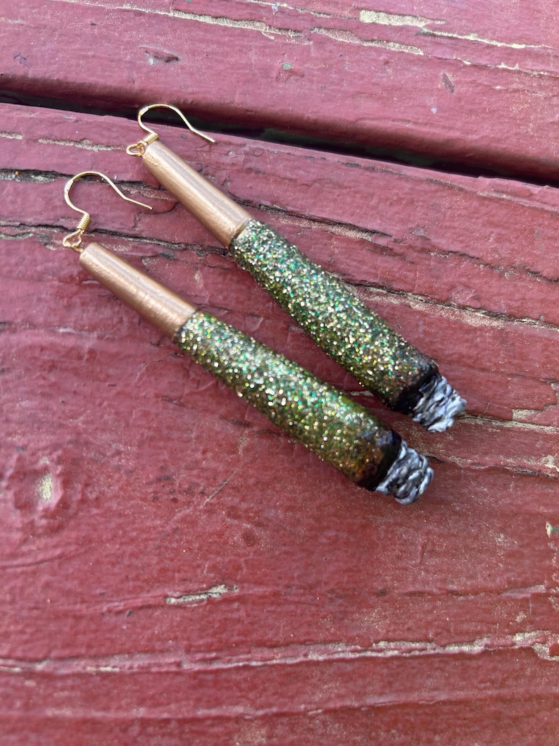 May include: A pair of handmade earrings designed to resemble rolled cigarettes. The earrings feature gold-colored metal tubes, glitter-covered bodies in shades of green and gold, and white and black accents at the ends.