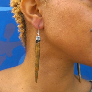 May include: A pair of long, wooden earrings with a silver hook and a small, round, silver bead at the top. The earrings are a light brown color with a darker brown stripe running down the center.