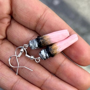 May include: A pair of novelty earrings featuring pink, cone-shaped charms designed to resemble lit cigarettes. The charms have a gradient of colors, including pink, brown, black, and white. The earrings are attached to silver-colored fish hook ear wires.