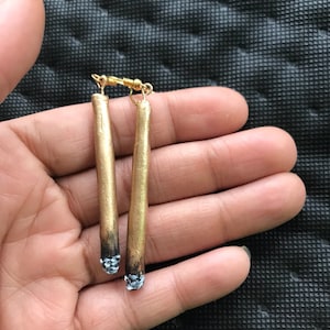 May include: Gold-colored earrings shaped like cigarettes. The earrings have a gold hook closure and feature a dark, burnt end with a white and blue ash design. The earrings are held in a hand against a dark background.