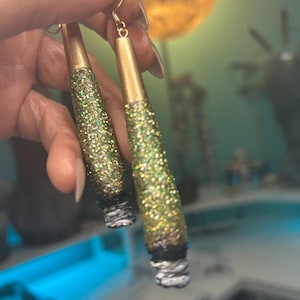 May include: A pair of handmade earrings with a gold-colored top and a glittery green and gold body. The bottom of the earrings have a white and gray design. The earrings are hanging from gold-colored hooks.