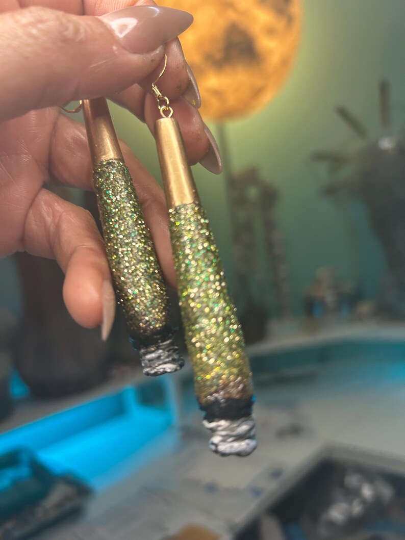May include: A pair of handmade earrings with a gold-colored top and a glittery green and gold body. The bottom of the earrings have a white and gray design. The earrings are hanging from gold-colored hooks.