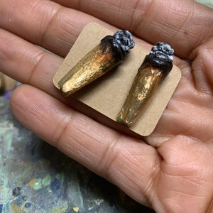 May include: A pair of gold-colored earrings shaped like rolled cigarettes with a dark, textured end. The earrings are displayed on a small, square, tan-colored card, held in a hand.