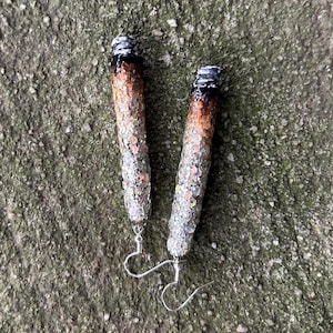 May include: A pair of drop earrings shaped like cylindrical objects with a gradient finish. The top is black, transitioning into a brown midsection, and ending in a silver, glittery base. Each earring features a silver-toned metal hook for attachment.