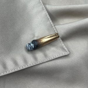May include: A small, three-dimensional pin shaped like a lit cigarette. The object features a metallic gold filter section, a dark brown burnt middle portion, and a textured, light gray ash tip. The pin is attached to a beige, smooth fabric garment.