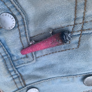May include: A pink and gray sparkly pin shaped like a joint, on a light blue denim jacket.