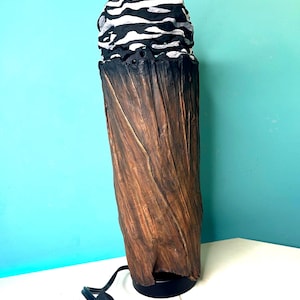 May include: A cylindrical table lamp with a textured brown base resembling tree bark. The top section features a contrasting black and white abstract pattern. A black power cord with an inline switch extends from the base of the lamp.