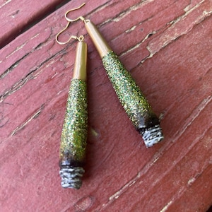 May include: A pair of handmade earrings with a unique design. The earrings feature a gold-colored top, a green glitter-covered center, and a white and gray textured bottom. The earrings hang from gold-colored ear wires.