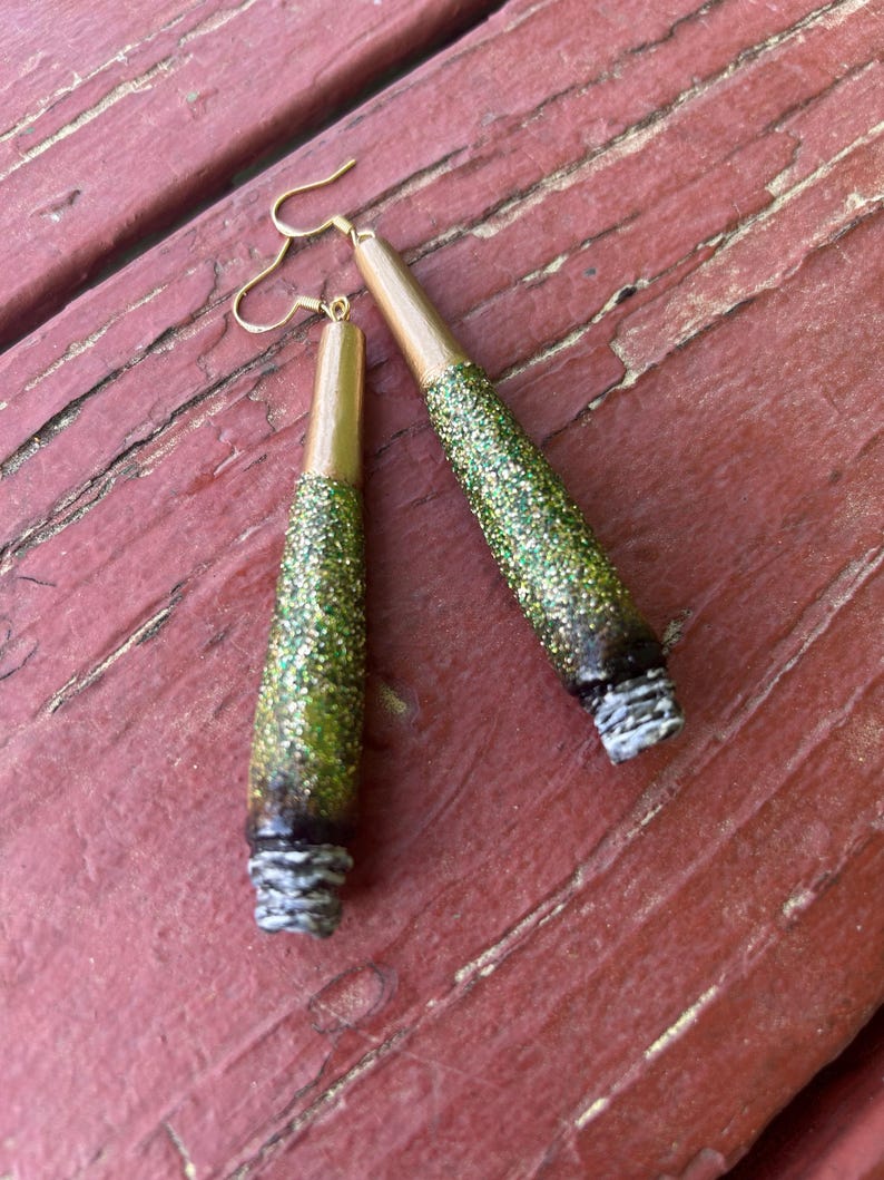 May include: A pair of handmade earrings with a unique design. The earrings feature a gold-colored top, a green glitter-covered center, and a white and gray textured bottom. The earrings hang from gold-colored ear wires.