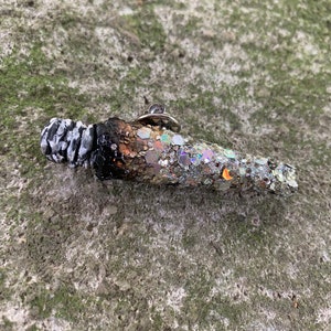 May include: A silver glitter pin shaped like a burnt out cigarette. The pin has a black and white tip and a silver glitter body.