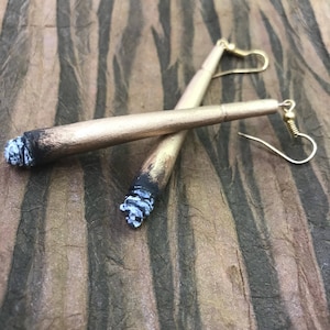 May include: A pair of gold-colored earrings shaped like cigarettes with white painted details. The earrings are hanging from gold hooks.