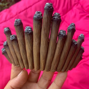 May include: A crown made of brown, lit cigarettes with white ash at the ends. The cigarettes are arranged in a circular pattern, set against a bright pink background.