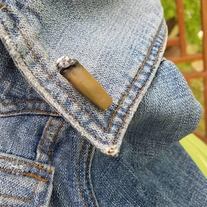 May include: A close-up of a denim jacket with a brown and white pin on the collar.