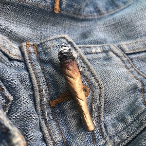 Puede incluir: Primer plano de un porro parcialmente fumado, con un exterior de papel marrón y una punta de ceniza blanca, metido en el bolsillo de una chaqueta vaquera azul.