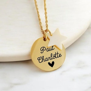 Op de afbeelding: Goudkleurige ketting met een ronde hanger en een sterbedel. De hanger heeft de namen "Paul" en "Charlotte" in script, met een klein hartje eronder. De sterbedel is wit.