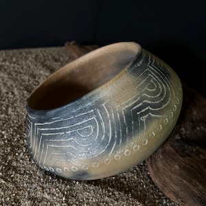 May include: A rounded, hand-formed clay bowl featuring a matte, tan-colored surface with dark charcoal-gray smoke-fired gradients. The exterior is decorated with incised geometric patterns consisting of concentric lines and a row of small circles.