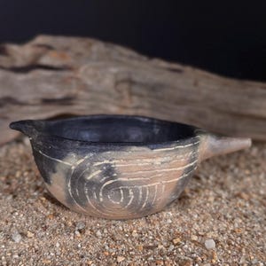 May include: A small, round, hand-formed clay bowl with a dark, charred black interior and a light tan exterior. The exterior features a carved spiral pattern and a small, pointed spout extending from one side, creating a unique, rustic vessel.