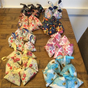 May include: A collection of 20 handmade fabric triangular sachets in various colours and patterns, including floral, bee, and geometric designs. The sachets are arranged in rows on a wooden table.