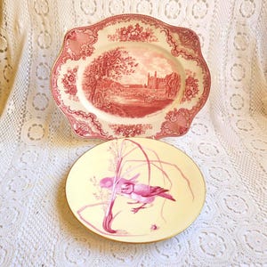May include: Two decorative plates. The top plate is a scalloped-edge plate with a red and white landscape design. The bottom plate is round with a yellow background and a pink bird illustration with a gold rim.