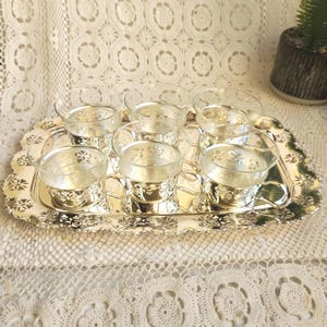 May include: A silver-toned metal serving tray with a scalloped edge and intricate cut-out patterns, holding six clear glass cups. Each glass cup is encased in a matching silver-toned metal holder featuring a decorative floral cut-out design.
