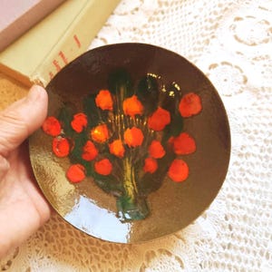 May include: A small, round, brown dish with a floral design. The design features orange and red flower-like shapes with green leaves. The dish is held by a hand, and the background is a white, lacy fabric.