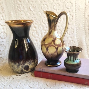 May include: Three vintage ceramic vases. The largest is dark brown with a gold rim and a textured, off-white base. The middle vase is tan with a brown pattern and a gold rim. The smallest vase is brown and green. All are on a white surface.