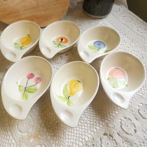 May include: Set of six off-white ceramic bowls with a unique handle design. Each bowl features a different hand-painted fruit design in the center, including a lemon, orange, blueberry, cherries, pear, and a peach. The bowls are arranged on a lace tablecloth.