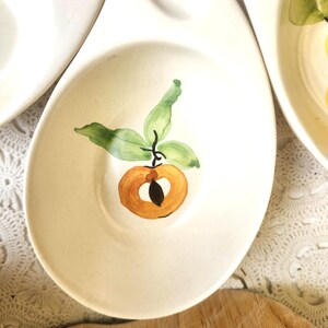 May include: Off-white ceramic serving dish with a hand-painted peach design. The peach is orange with a black center, and green leaves. The dish has a unique shape with a handle. The design is centered on the dish.