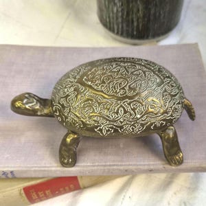 May include: A brass turtle figurine featuring an intricate, engraved pattern across its shell. The metal surface has a polished, reflective finish with dark recessed lines highlighting the detailed, swirling decorative motifs on the back.