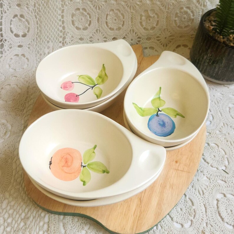 May include: Three off-white ceramic bowls with a handle, each decorated with a different fruit: cherries, a blueberry, and a peach. The bowls are arranged on a wooden cutting board.
