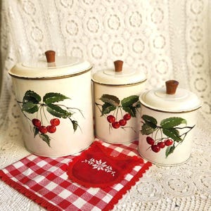 May include: A set of three vintage cream-colored kitchen canisters with cherry branch illustrations. Each canister has a wooden knob on the lid and a gold rim. A red and white checkered napkin and a red bib with white embroidery are in the foreground.