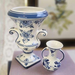 May include: A white ceramic mug with a black handle and the text "But First, Coffee" in a bold, black font. The mug is set against a blurred background. The mug appears to be approximately 10 cm tall.