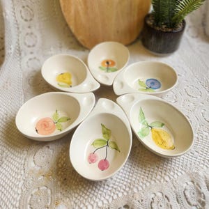 May include: Six off-white ceramic serving dishes, each with a unique hand-painted fruit or floral design. The designs include a yellow lemon, pink cherries, a blue blueberry, and a yellow flower. Each dish has a handle.