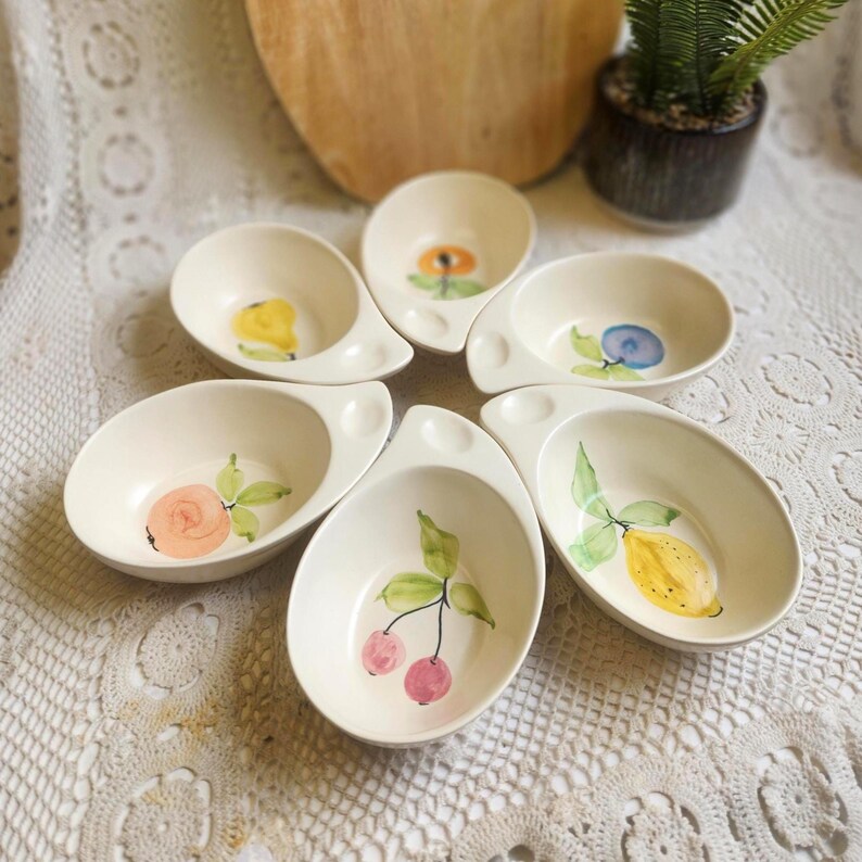 May include: Six off-white ceramic serving dishes, each with a unique hand-painted fruit or floral design. The designs include a yellow lemon, pink cherries, a blue blueberry, and a yellow flower. Each dish has a handle.