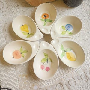 May include: Six off-white ceramic serving bowls, each with a unique hand-painted fruit design. The fruits include a pear, peach, blueberry, rose, cherries, and a lemon. Each bowl has a small handle.