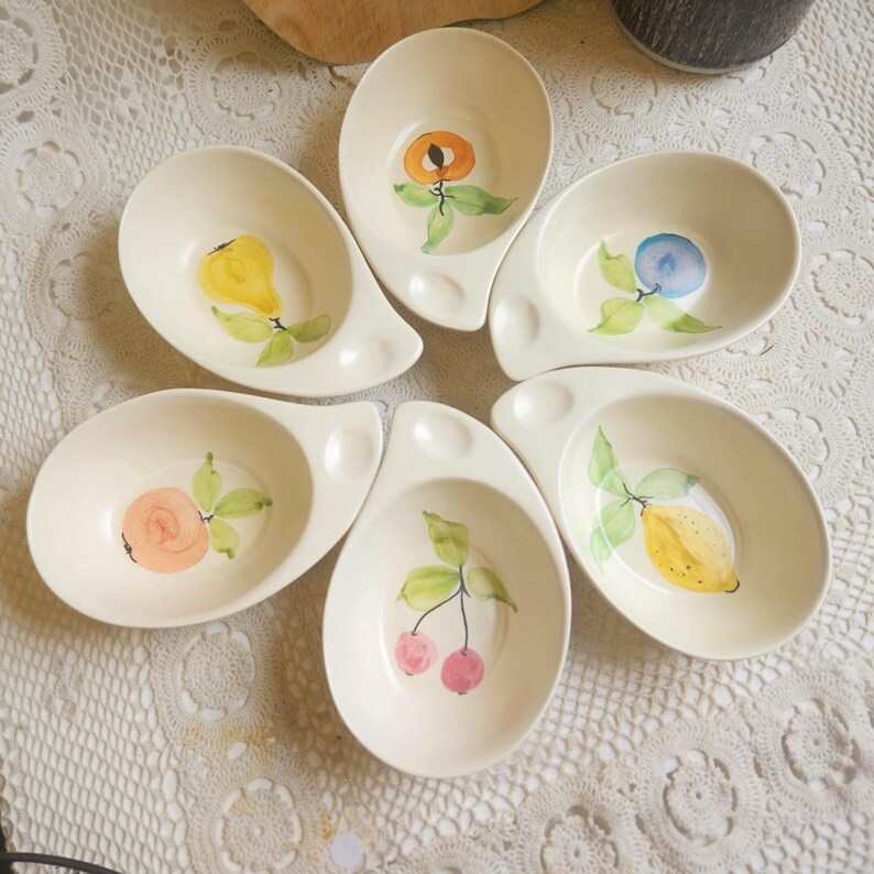 May include: Six off-white ceramic serving bowls, each with a unique hand-painted fruit design. The fruits include a pear, peach, blueberry, rose, cherries, and a lemon. Each bowl has a small handle.