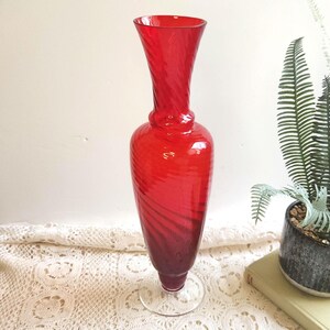 May include: A tall, vibrant red glass vase featuring a distinct spiral or optic twist pattern throughout its body. The vase has a slender neck that flares slightly at the top rim and sits on a clear, circular glass base, showcasing a translucent finish.