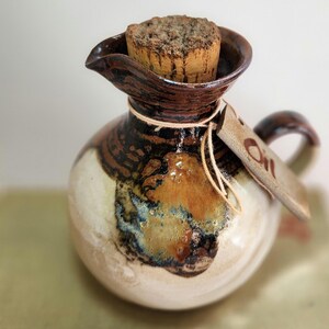 May include: A handmade ceramic oil cruet with a cork stopper. The cruet has a round body, spout, and handle. The colour scheme includes brown, cream, and blue tones. A tag with the word "Oil" is attached with twine.
