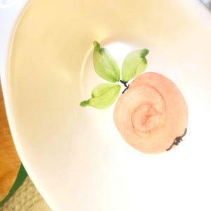 May include: A white ceramic plate with a painted peach design. The peach is a light orange color with a spiral pattern, and green leaves. The plate has a slightly off-white color.