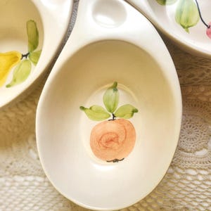 May include: Off-white ceramic serving dish with a hand-painted peach design. The peach is a light orange color with green leaves. The dish has a unique shape with a handle and a small bowl.