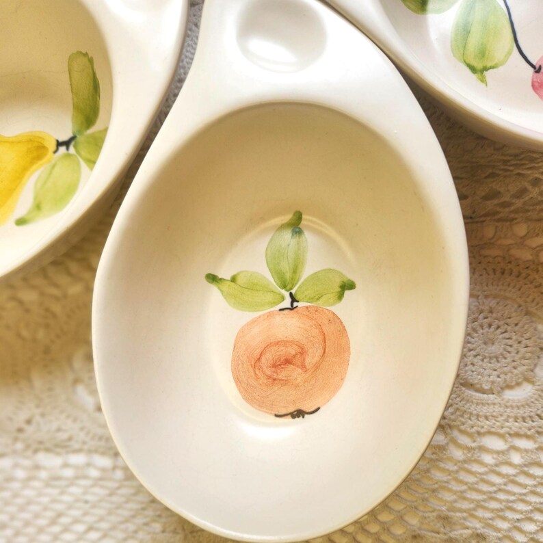 May include: Off-white ceramic serving dish with a hand-painted peach design. The peach is a light orange color with green leaves. The dish has a unique shape with a handle and a small bowl.
