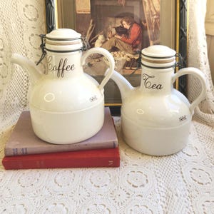 May include: Two white ceramic coffee and tea pots with black lettering. Each pot has a lid, handle, and spout. The coffee pot has the word "Coffee" and the tea pot has the word "Tea". The pots are sitting on top of books.