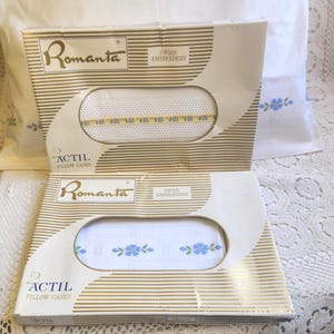 May include: Two boxes of Romanta Actil pillowcases. The boxes are white and gold striped with the words "Swiss Embroidery" printed on them. The pillowcases are white with blue floral embroidery.
