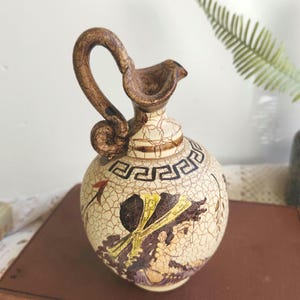 May include: A ceramic jug with a brown handle and spout, featuring a cream-colored body with a cracked glaze effect. The jug is adorned with black geometric patterns and illustrations in shades of brown, yellow, and purple, reminiscent of ancient Greek art.